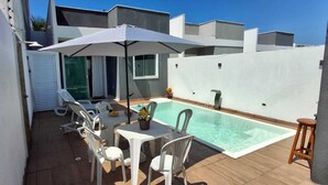 A modern outdoor pool area with a white umbrella, white plastic chairs, and a small table with a vase of flowers.