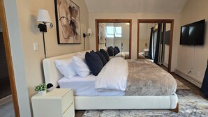 Ein modernes Hotelzimmer mit einem großen Bett, einem Flachbildfernseher, einem Nachttisch mit Lampe und Blick ins Freie durch ein großes Fenster.
