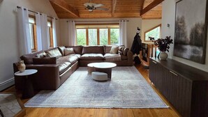 Ein geräumiges Wohnzimmer mit einer großen, L-förmigen Couch, einem runden Couchtisch und einem Fernseher auf einem Holzschrank. Große Fenster sorgen für natürliches Licht und Blick auf die grüne Umgebung.
