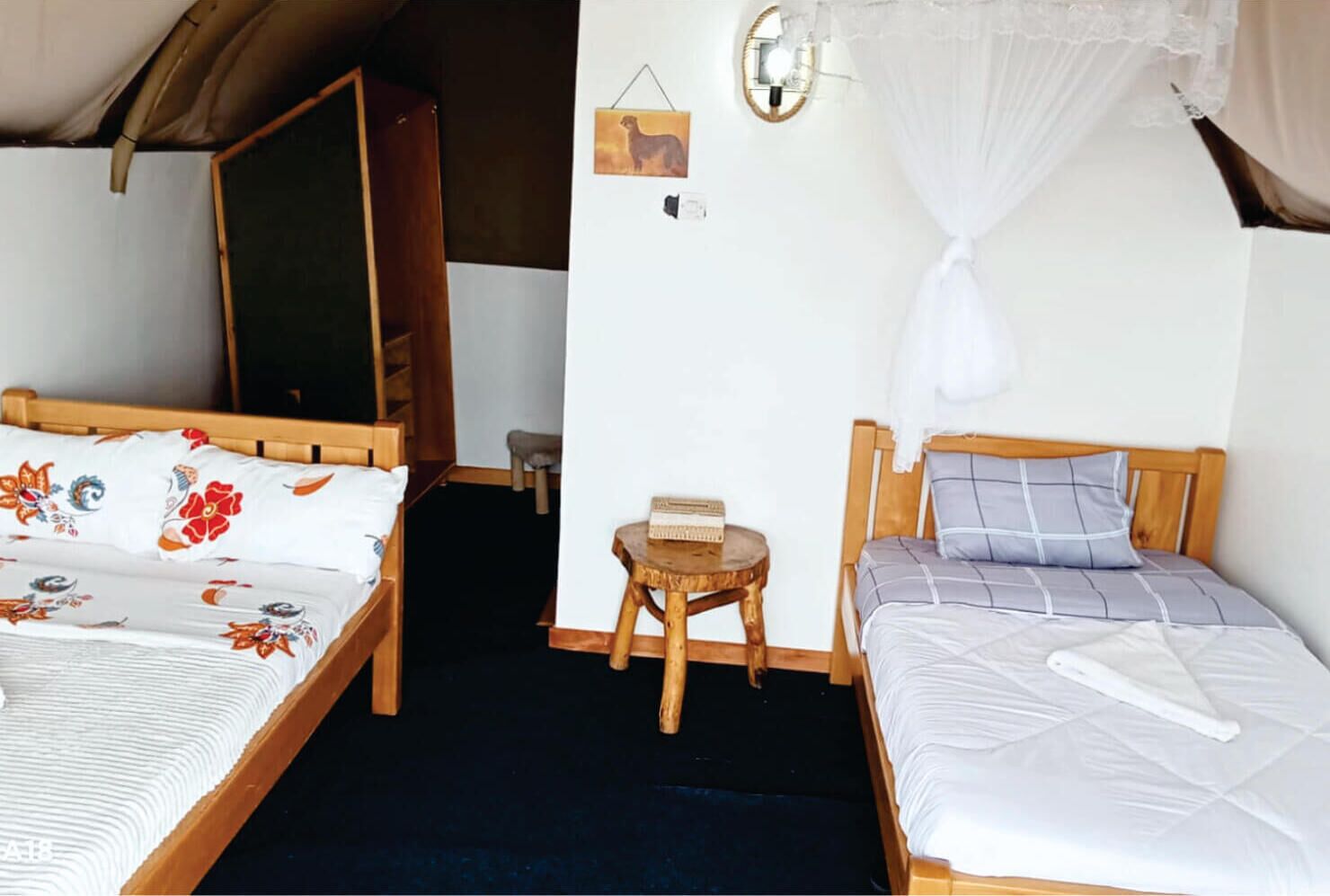 A room with two wooden beds, one with floral bedding and the other with a checkered pattern. There is a small wooden table with a book and a wall-mounted light fixture.