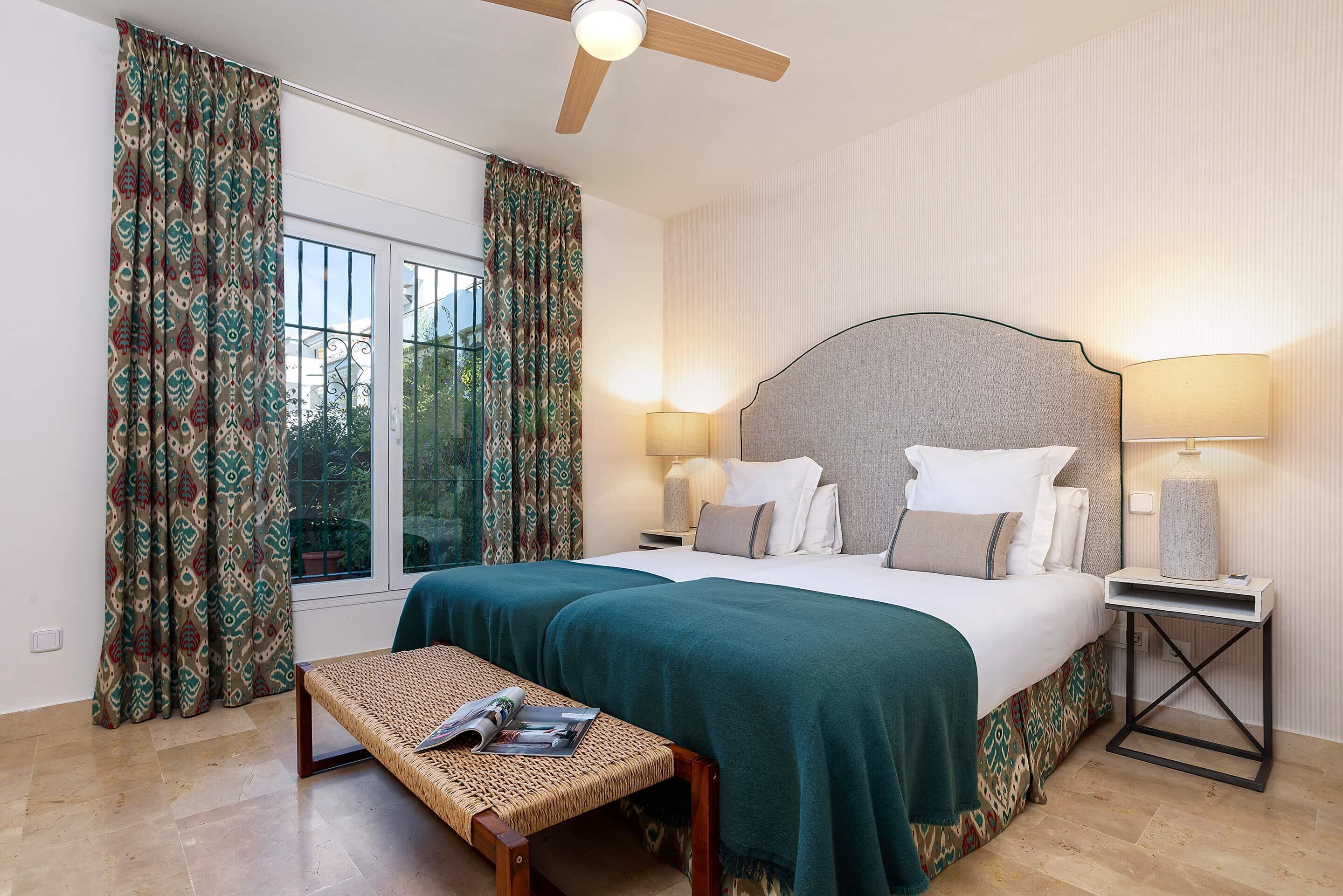 Une chambre d’hôtel avec un grand lit, deux lampes de chevet, un banc avec un magazine et une fenêtre avec des rideaux.