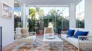 Um pátio ensolarado com um sofá branco, cadeiras de vime e uma mesa de centro.