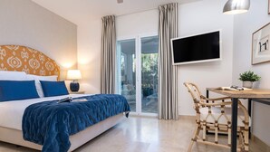 Quarto de hotel com uma cama, uma escrivaninha, uma cadeira, uma televisão e uma porta de vidro deslizante para o exterior.