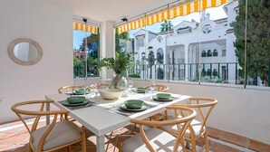 Uma área de refeições com uma mesa branca posta para quatro pessoas, rodeada por cadeiras de vime com almofadas. Uma janela grande com vista para um edifício branco e vegetação é visível.
