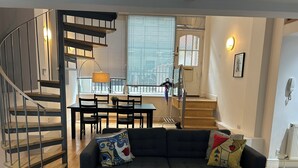 Ein modernes Wohnzimmer mit grauem Sofa, einer Holztreppe, einem Essbereich und großen Fenstern.