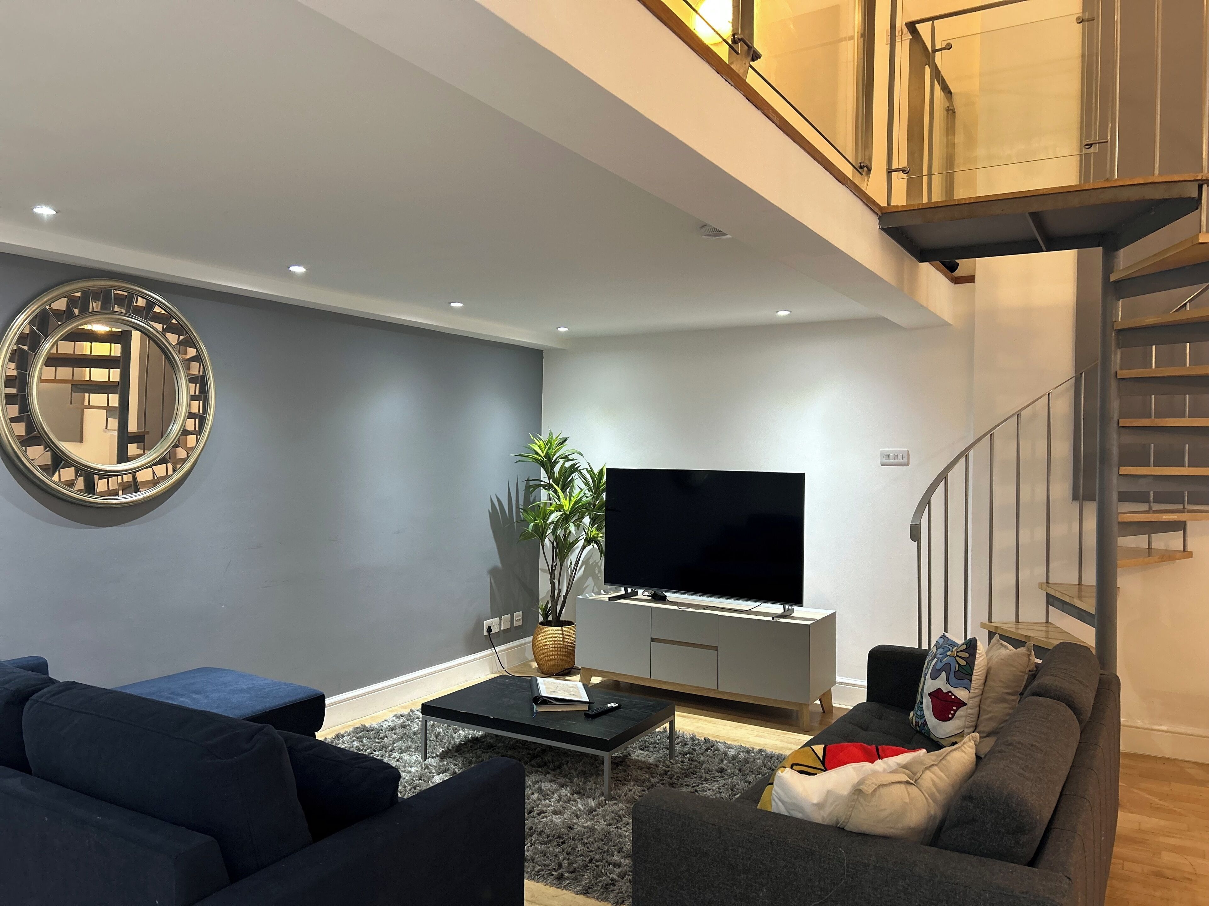 A modern living room with a flat-screen TV, grey sofas, a coffee table, and a staircase.