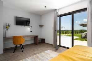 Une chambre d’hôtel moderne dotée d’un bureau en bois, d’un téléviseur à écran plat, d’un fauteuil confortable, d’un lit et d’une porte-fenêtre coulissante donnant sur un balcon avec vue sur une piscine et un espace verdoyant.