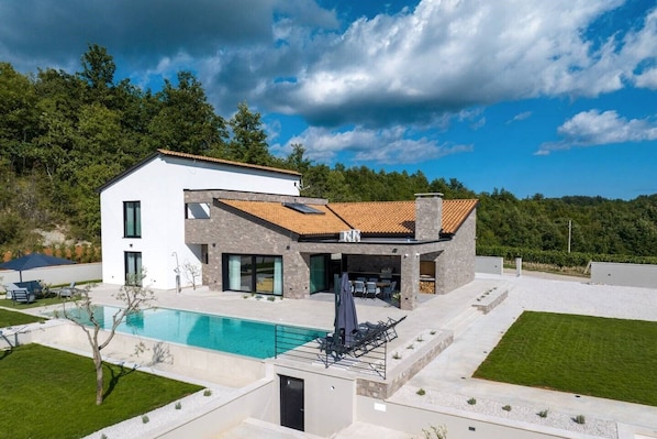 Une maison moderne à deux étages, dotée d’une piscine, d’un espace de détente extérieur et offrant une vue sur la verdure environnante.