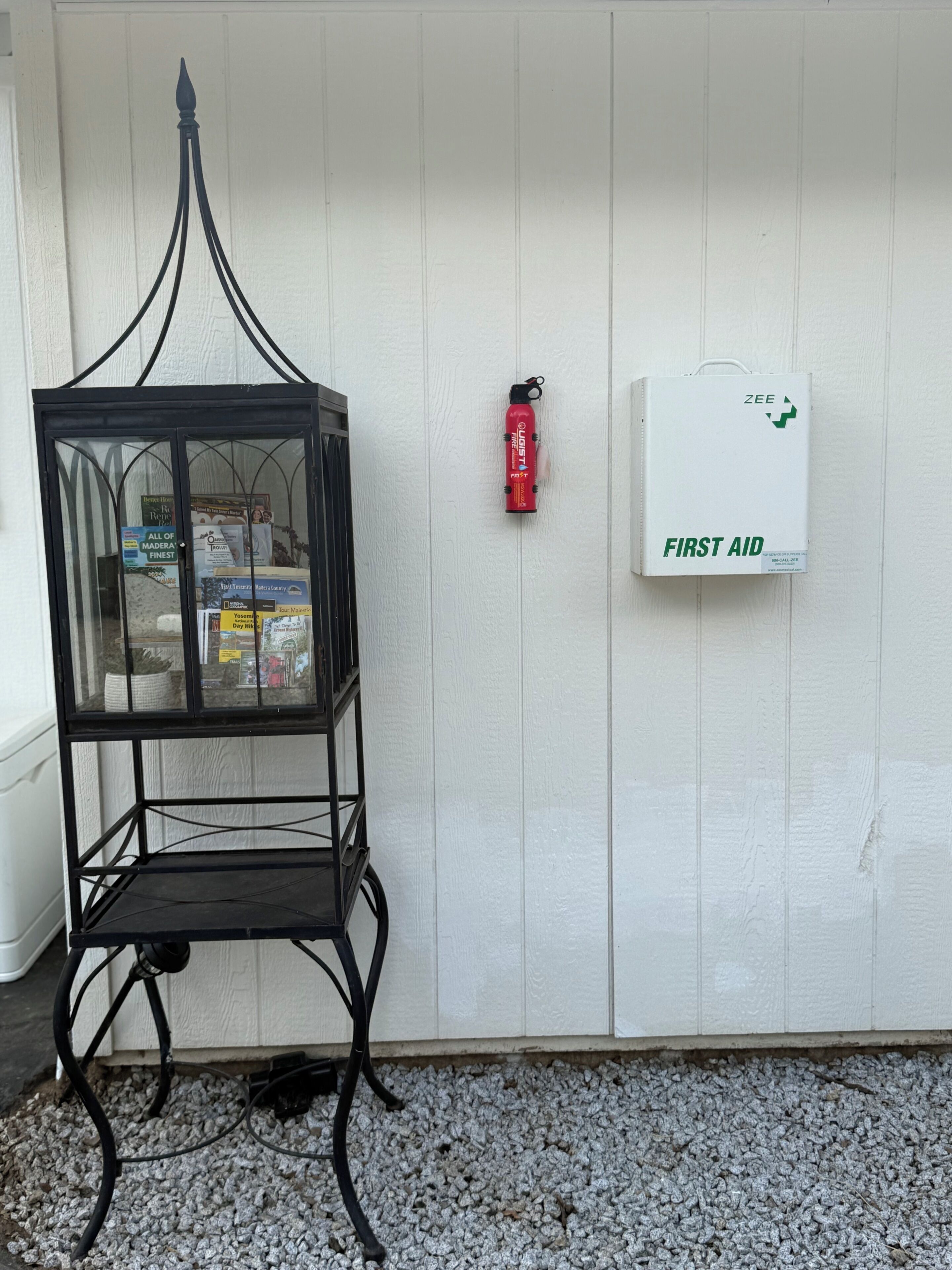 Ein schwarzer Metallstuhl mit Glaseinsatz, ein roter Feuerlöscher und eine weiße Schachtel mit der Aufschrift 'FIRST AID' an einer weißen Wand angebracht.