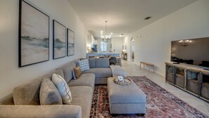 A modern living room with a sectional sofa, ottoman, and a large flat-screen TV. There is a kitchen in the background with a dining area. - Modern Heritage Landing Condo – Resort Lifestyle (Punta Gorda)