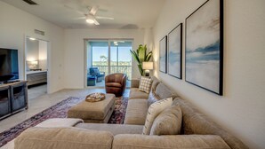 A spacious living room with a large beige sectional sofa, a flat-screen TV, a ceiling fan, and a large window offering a view of the outdoors. - Modern Heritage Landing Condo – Resort Lifestyle (Punta Gorda)