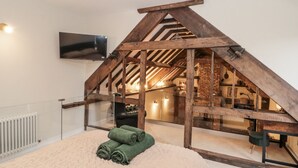 Dormitorio tipo loft con vigas de madera a la vista, un televisor montado en la pared y vistas hacia una zona de cocina.