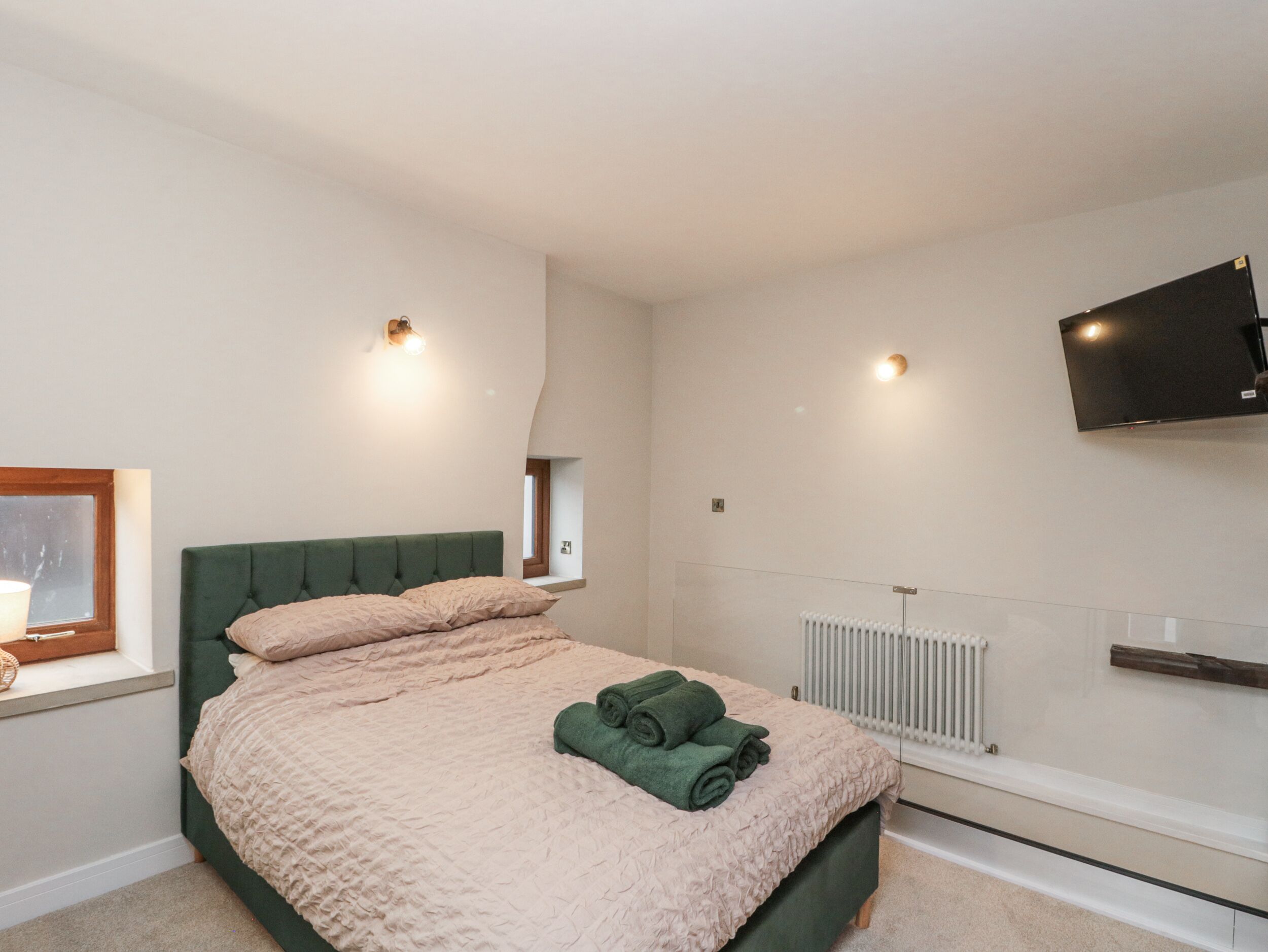 A bedroom with a green upholstered headboard, a pink textured bedspread, and a rolled green towel on the bed. There is a window with a wooden frame, a wall-mounted TV, and a white radiator.