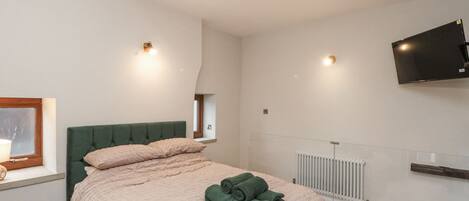A bedroom with a green upholstered headboard, a pink textured bedspread, and a rolled green towel on the bed. There is a window with a wooden frame, a wall-mounted TV, and a white radiator.