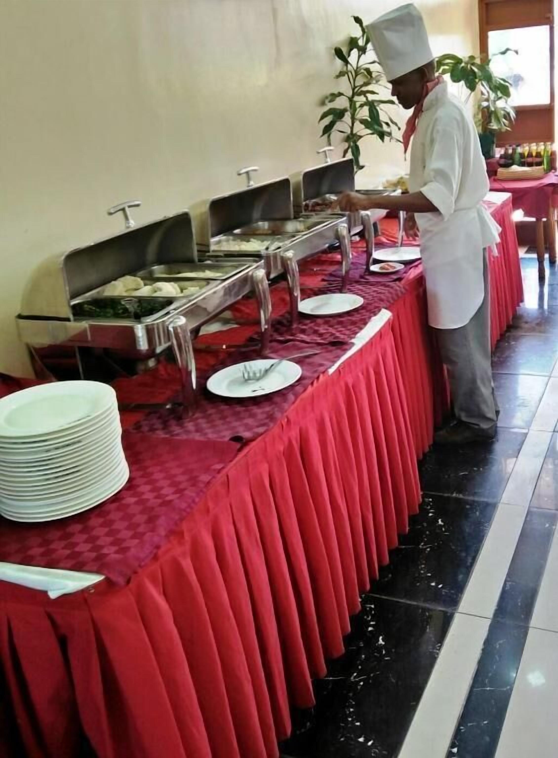 En buffetopstilling med flere serveringsstationer, en stabel hvide tallerkener og en kok, der tilbereder mad.