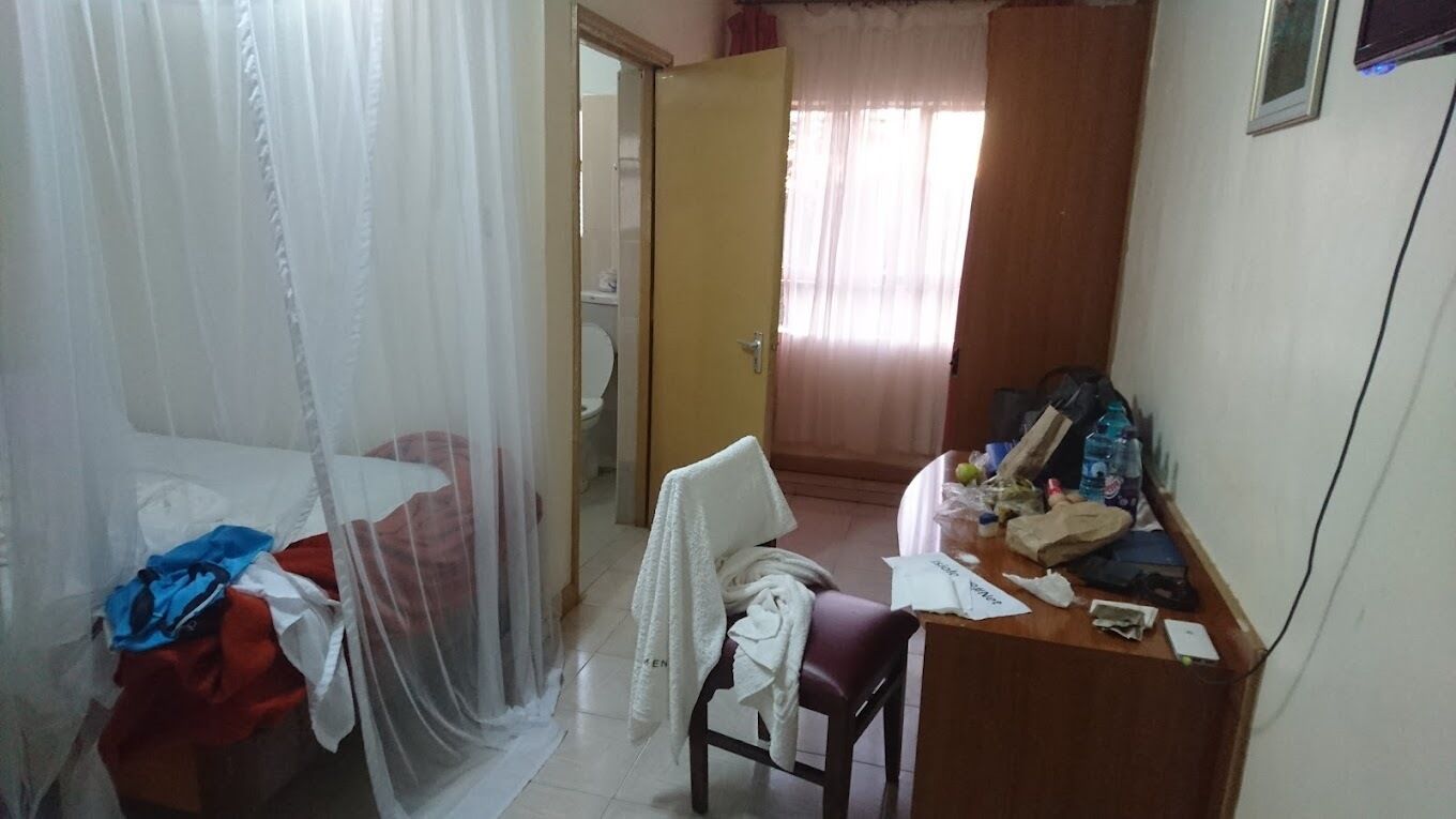 A small, dimly lit room with a bed covered in a mosquito net, a chair, a desk cluttered with items, and a door leading to a bathroom.