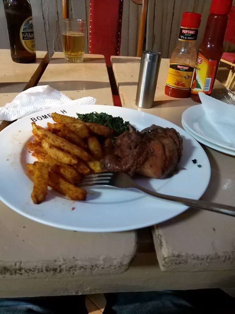 A plate with a piece of meat and fries, a bottle of beer, and condiments on a table.