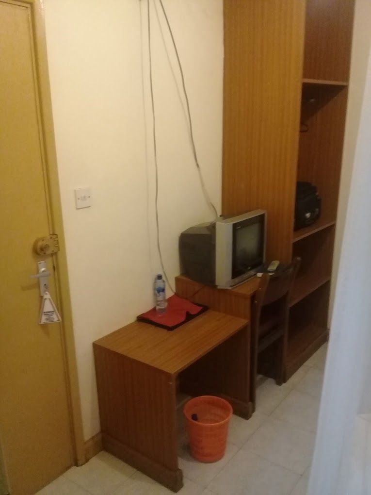 Une petite pièce avec un bureau en bois, une télévision, un dossier rouge et un seau rouge posé sur le sol.