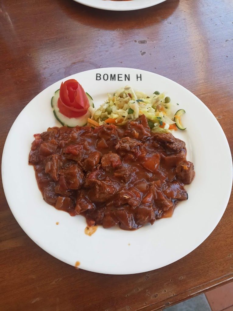 Une assiette de nourriture comprenant un plat de viande, une salade de légumes et une tranche de concombre garnie d’une tomate, le tout posé sur une table en bois.