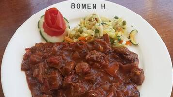 Une assiette de nourriture comprenant un plat de viande, une salade de légumes et une tranche de concombre garnie d’une tomate, le tout posé sur une table en bois.