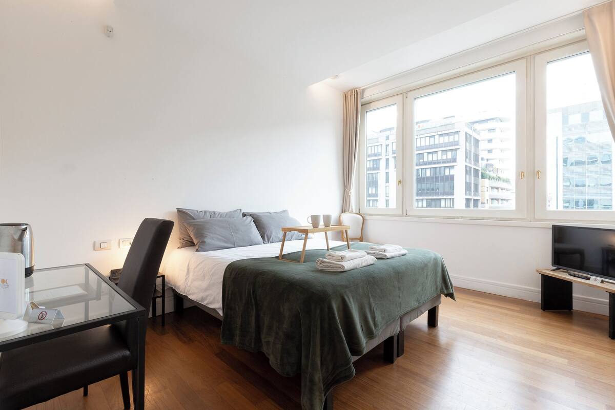 A modern hotel room with a bed, a glass table, a chair, a TV, and a window with a city view.