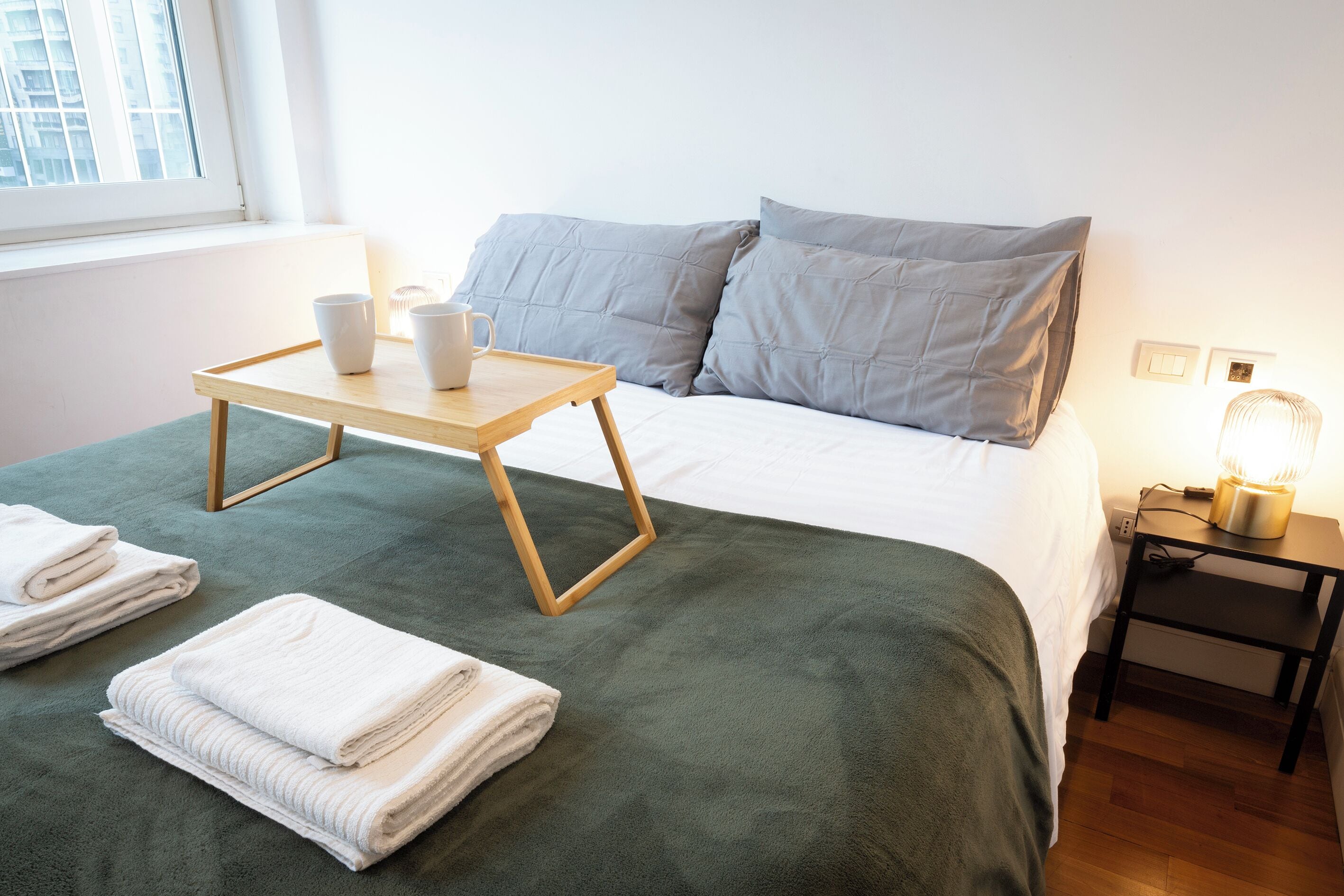 Ein ordentlich bezogenes Bett mit grüner Decke, ein Nachttisch mit Lampe und ein Tablett mit Tassen und einer Schüssel.