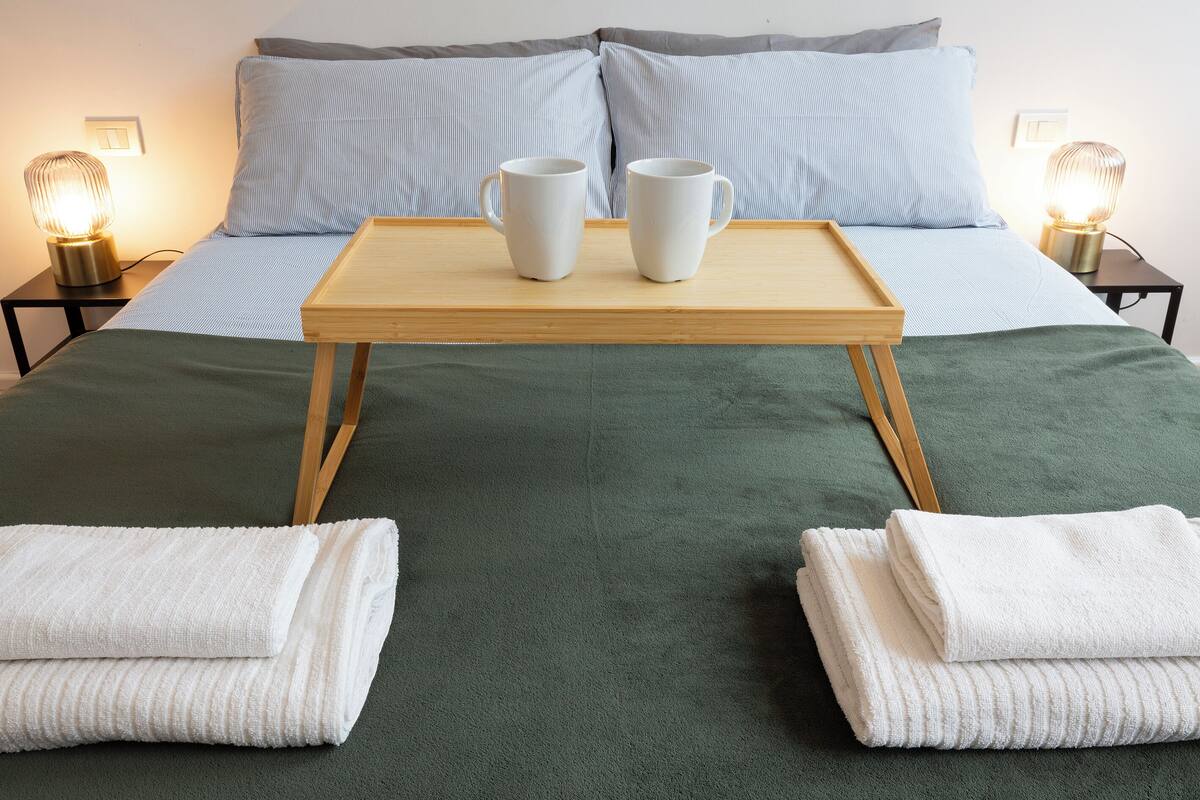 A neatly made bed with a green bedspread, two white towels at the foot of the bed, and a tray with two white cups on a wooden table in front of the bed.