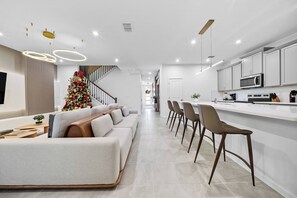 Unwind in a chic living area with a cozy L-shaped sofa and modern bar stools at the kitchen island. Unique light fixtures add warmth, enhancing the airy atmosphere and creating a welcoming vibe for your stay. - 6BR house with private pool in Solara Resort (Kissimmee)
