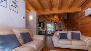 Ein gemütliches Wohnzimmer mit zwei beigen Sofas und dunklen Kissen, Holzbalkendecke und Blick auf die Küche und den Essbereich durch eine Glastür.