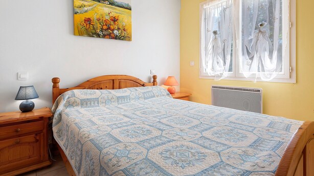 Een slaapkamer met een houten bed, een nachtkastje met een lamp, een radiator en een raam met dunne gordijnen.