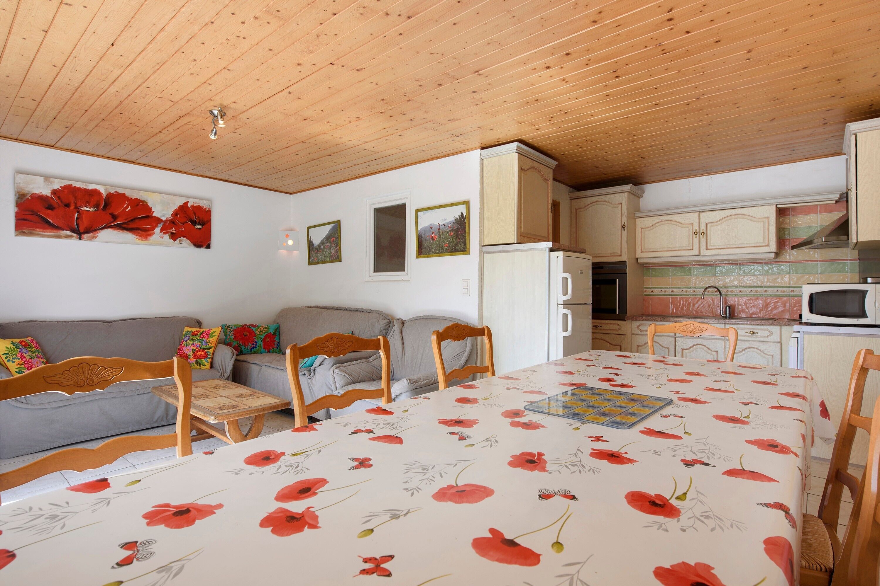 Ein gemütliches Zimmer mit Holzbalkendecke, einer Couch, einem Esstisch mit blumigem Tischlauf und einer Küchenzeile mit Schränken und Geräten.