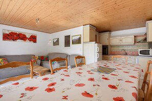 A cozy room with a wooden ceiling, a sofa, a dining table with a floral tablecloth, and a kitchen area with cabinets and appliances. - Apartment with Panoramic View in a Picturesque Mountain Village (Grust)