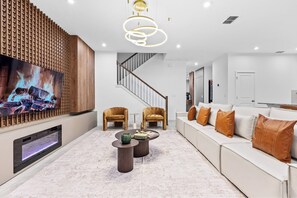 Ein modernes Wohnzimmer mit einem großen Flachbildfernseher, einer Holzwandverkleidung, einer Treppe, Ledersesseln, einem weißen Sofa mit orangefarbenen Kissen und einem Marmorboden.