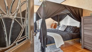 Un dormitorio con cama con dosel, suelo de madera, cómoda y ventana con cortinas.