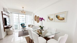 A modern living room with a glass dining table, white chairs, a blue armchair, a sofa, and a TV on a wooden shelf. - Beautiful apartment in Juan Dolio (Playa Juan Dolio)