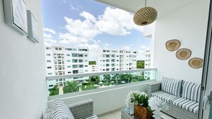 A balcony with wicker furniture, a hanging light fixture, and a view of white buildings and greenery. - Beautiful apartment in Juan Dolio (Playa Juan Dolio)