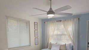 Un dormitorio con cama, ventilador de techo, ventana con persianas, una silla y una mesita de noche.