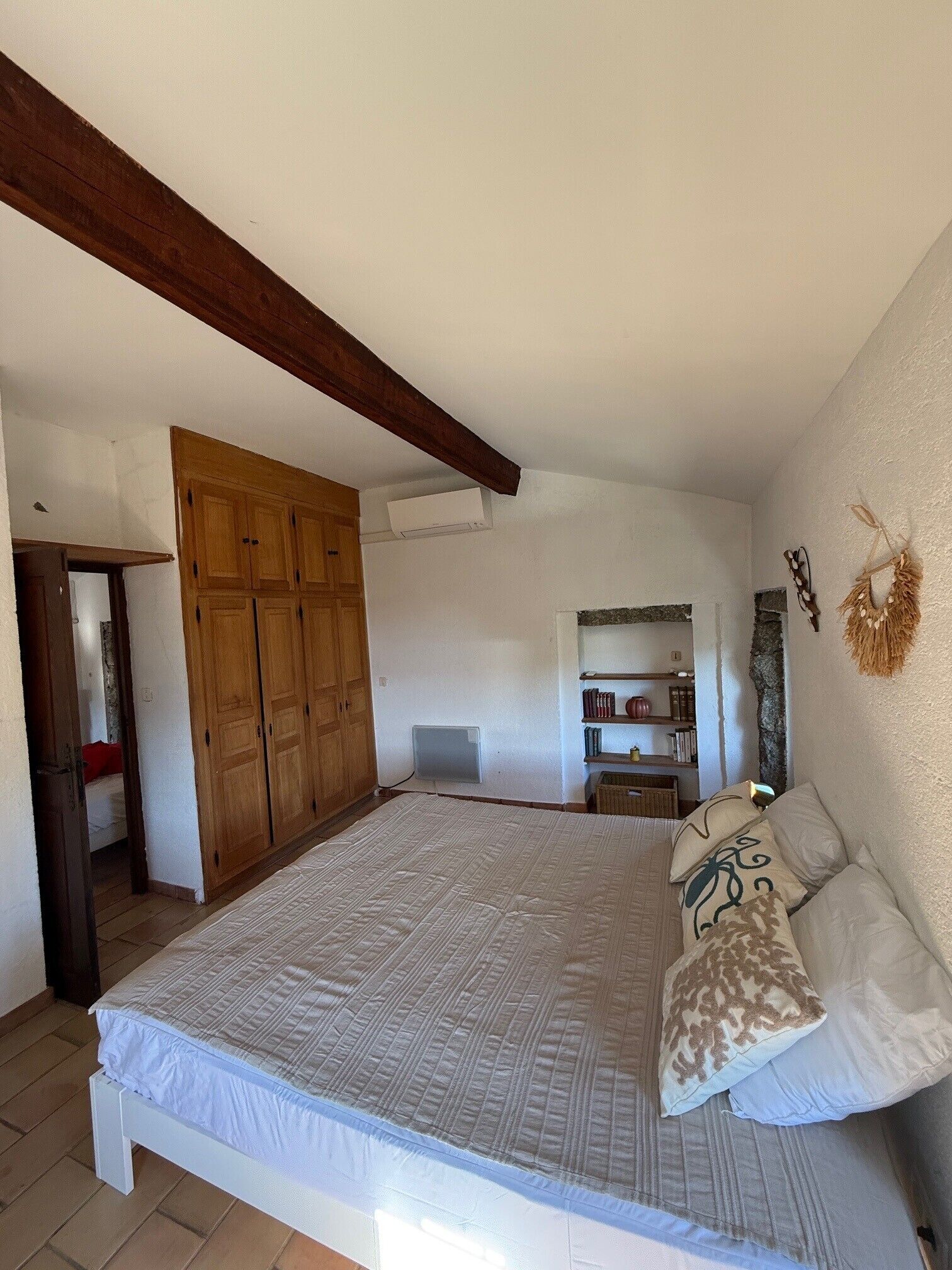 A bedroom with a large bed, wooden wardrobe, and a small shelf with books and decorative items.