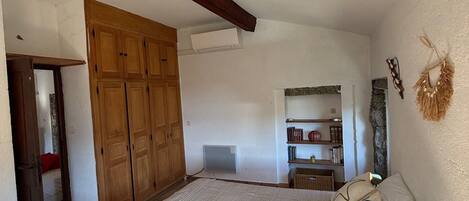 A bedroom with a large bed, wooden wardrobe, and a small shelf with books and decorative items.