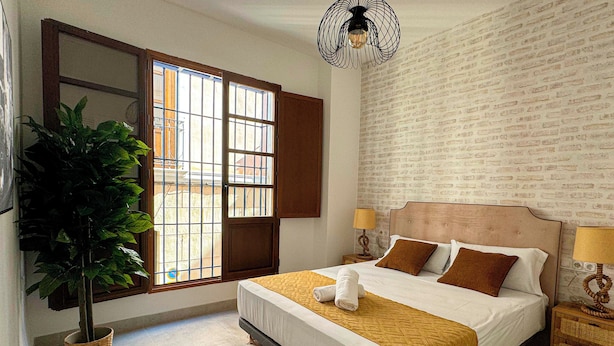 A hotel room with a large bed, a potted plant, a window with bars, and a brick-patterned wall. - Apartamentos Plaza San Agustin 3000 (Granada)