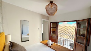 A hotel room with a bed, a mirror, a hanging light fixture, and a view of a tiled roof through an open door. - Apartamentos Plaza San Agustin 3000 (Granada)