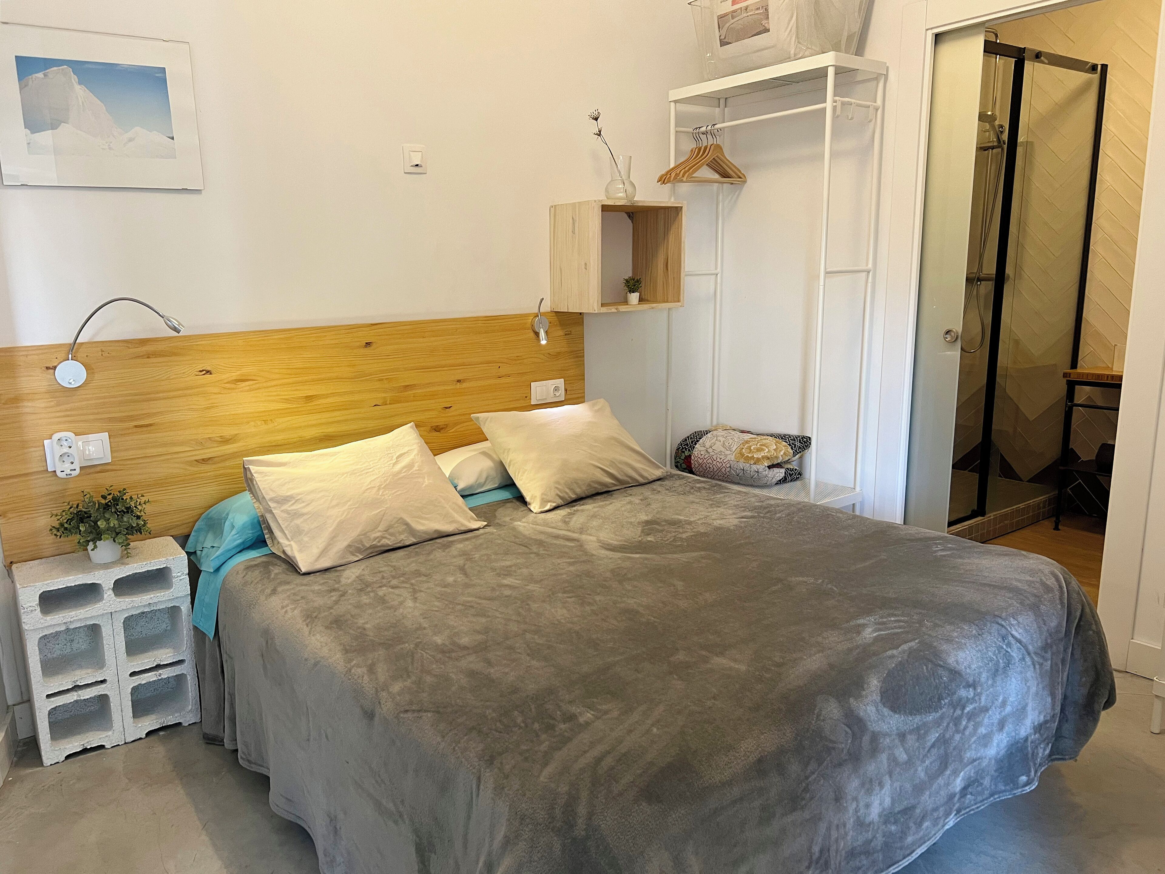 A bedroom with a wooden headboard, a bed with grey bedding, a bedside table with a plant, a small wooden shelf, and a walk-in closet with a hanging rack.