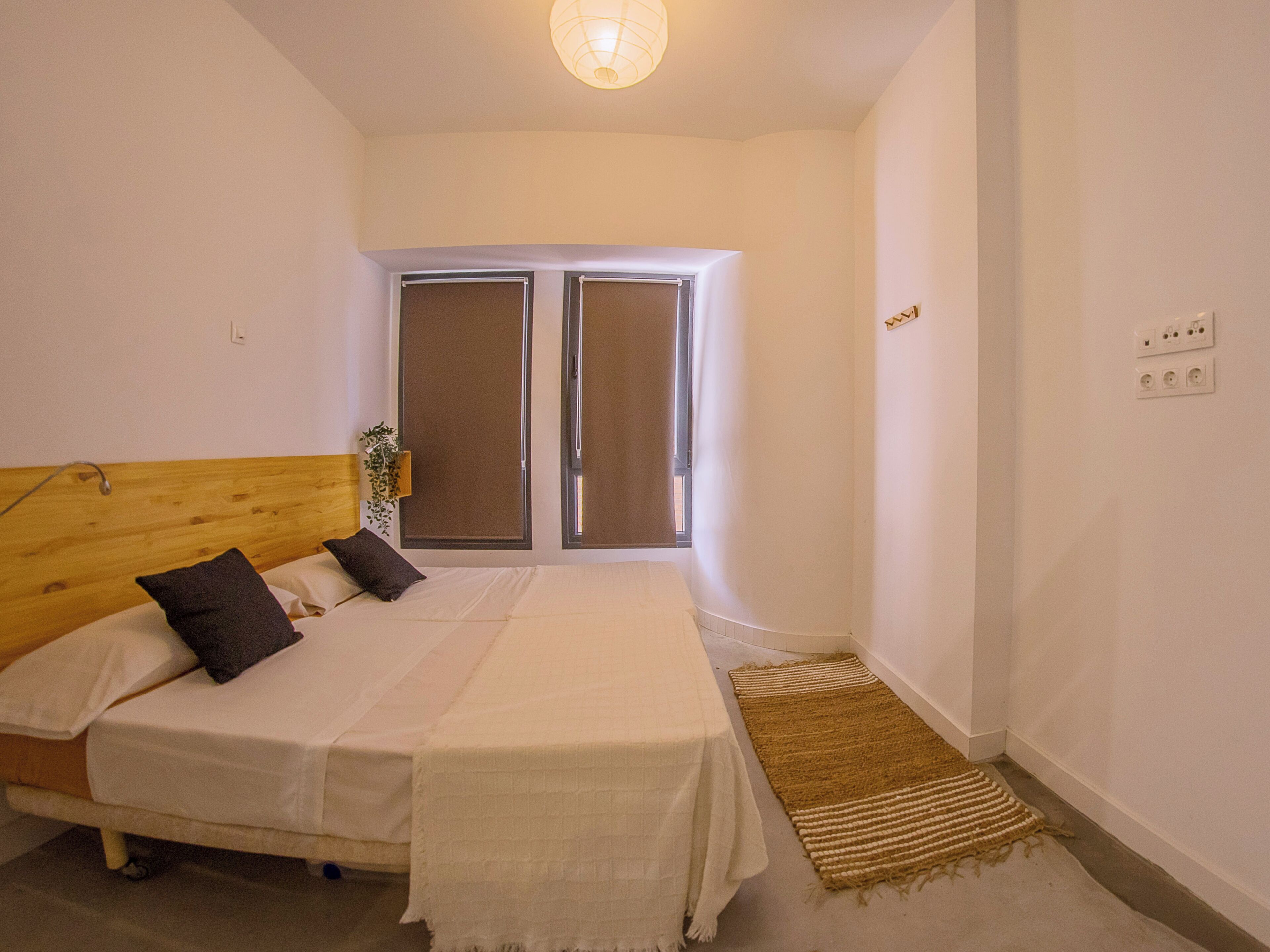 Un dormitorio con una cama, cabecera de madera, dos ventanas, una planta en la mesita de noche y una alfombra pequeña.