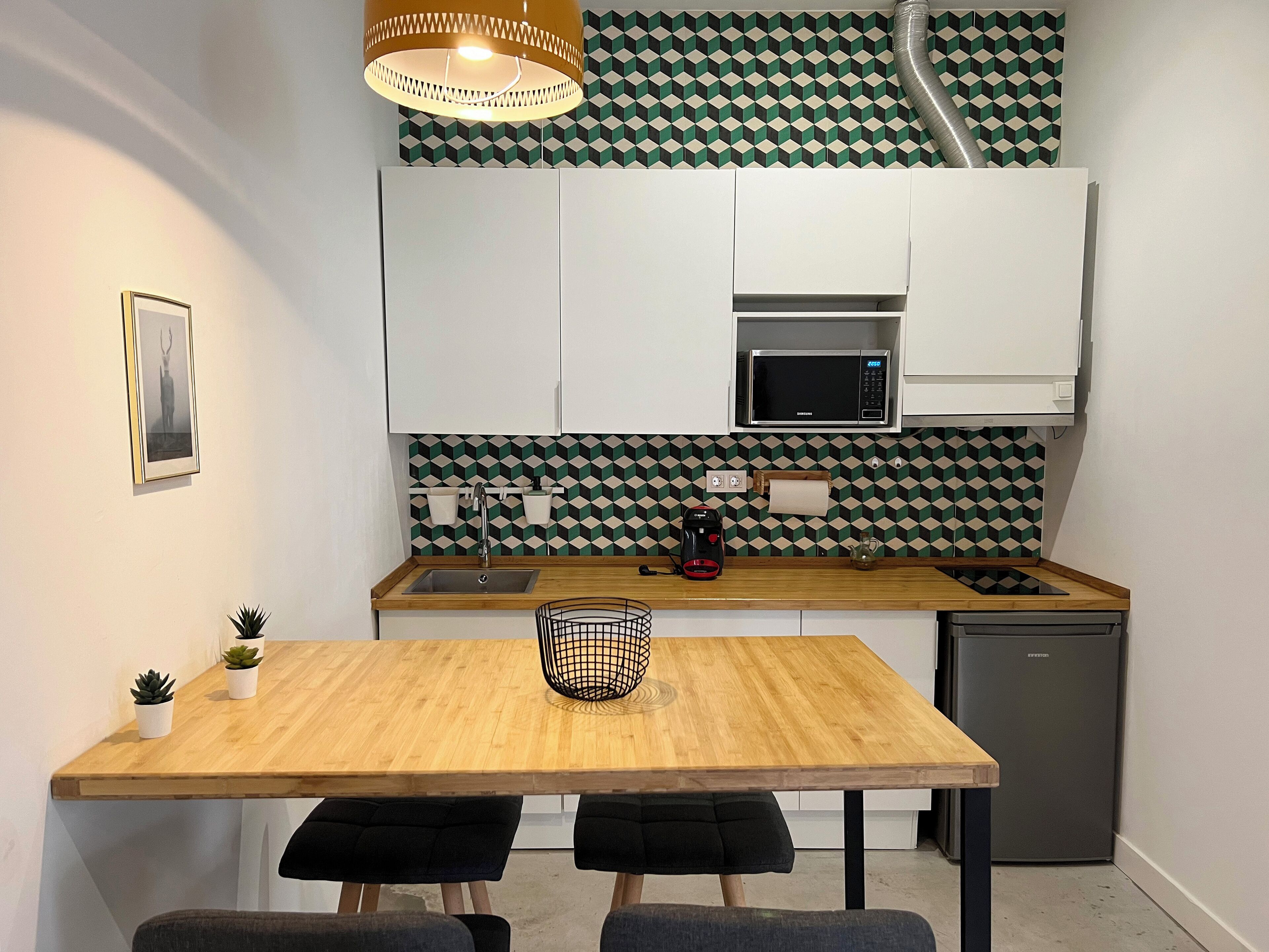 Une cuisine compacte avec une table en bois, des chaises noires rembourrées, un petit évier, un four à micro-ondes et un réfrigérateur.