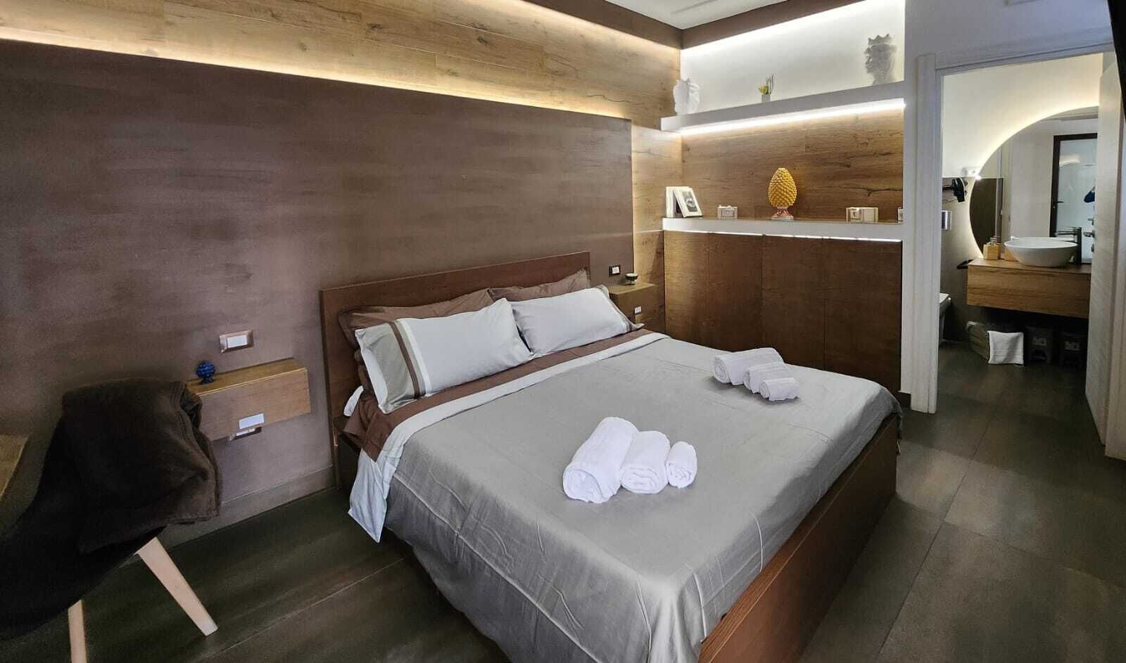 Ein modernes Hotelzimmer mit einem großen Bett, einem Holzkopfende, einem Nachttisch, einem Spiegel und einem durch einen Bogen sichtbaren Badezimmer.