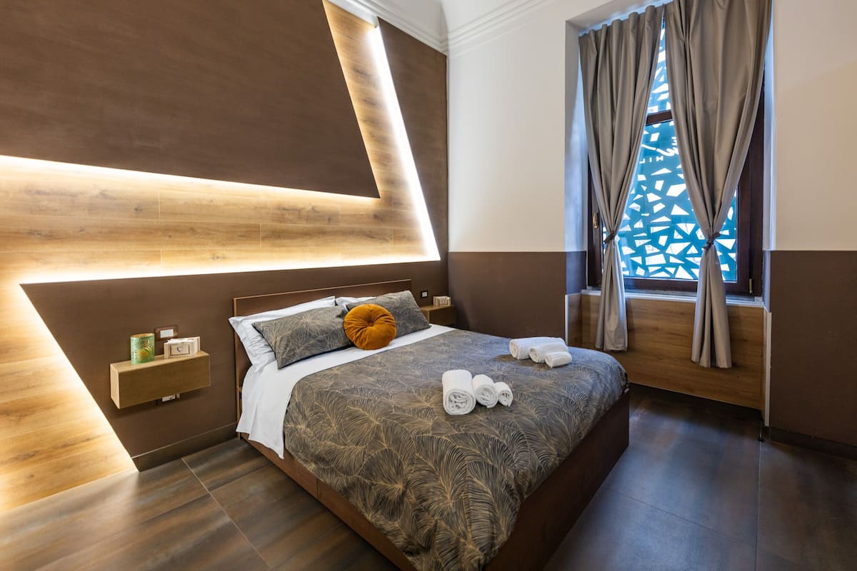 A modern bedroom with a wooden headboard, a large bed with a patterned bedspread, a bedside table with a lamp, and a window with curtains.