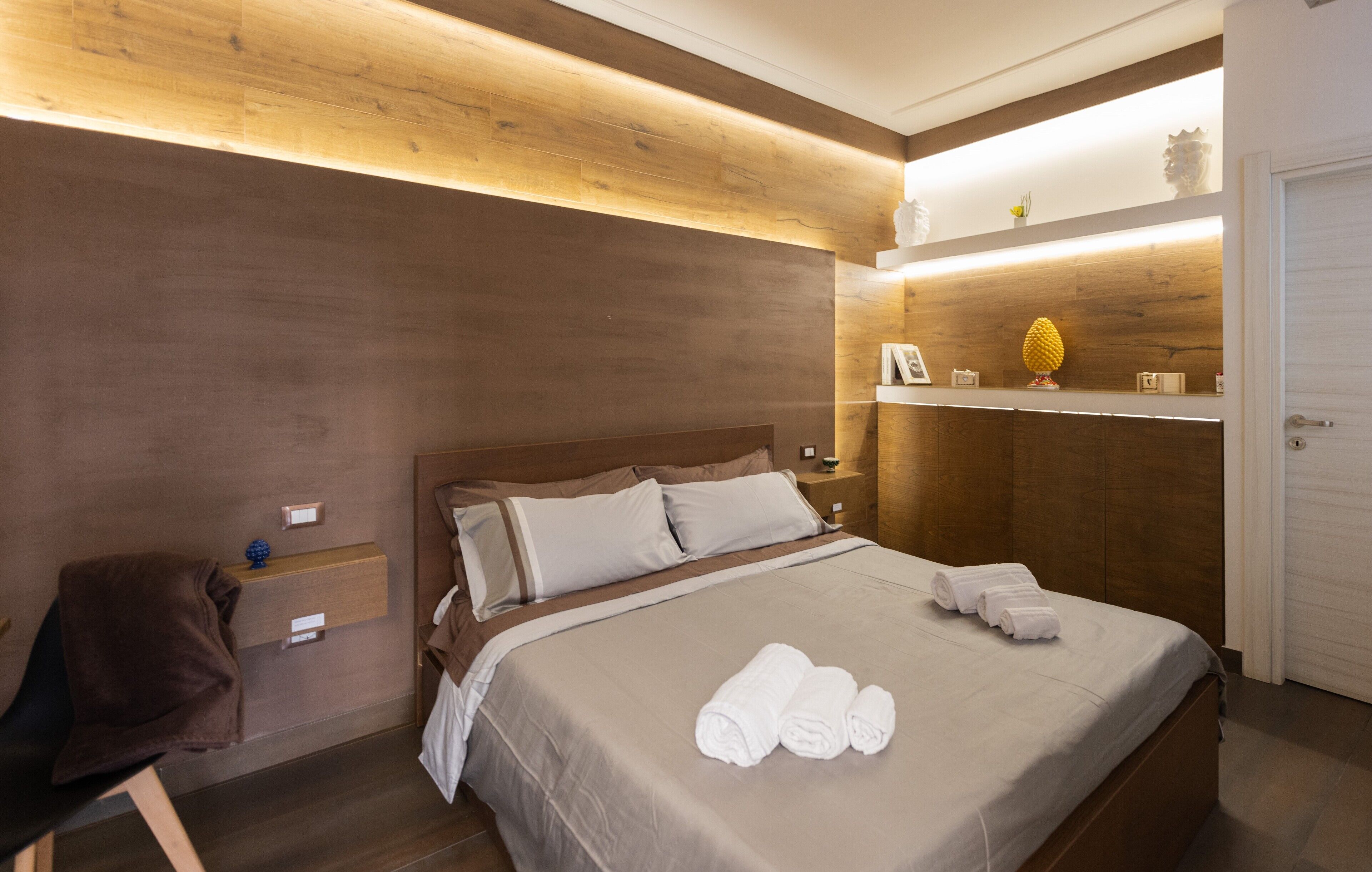 A modern hotel room with a wooden headboard, a bed with neatly folded towels, a nightstand, and a shelf with decorative items.