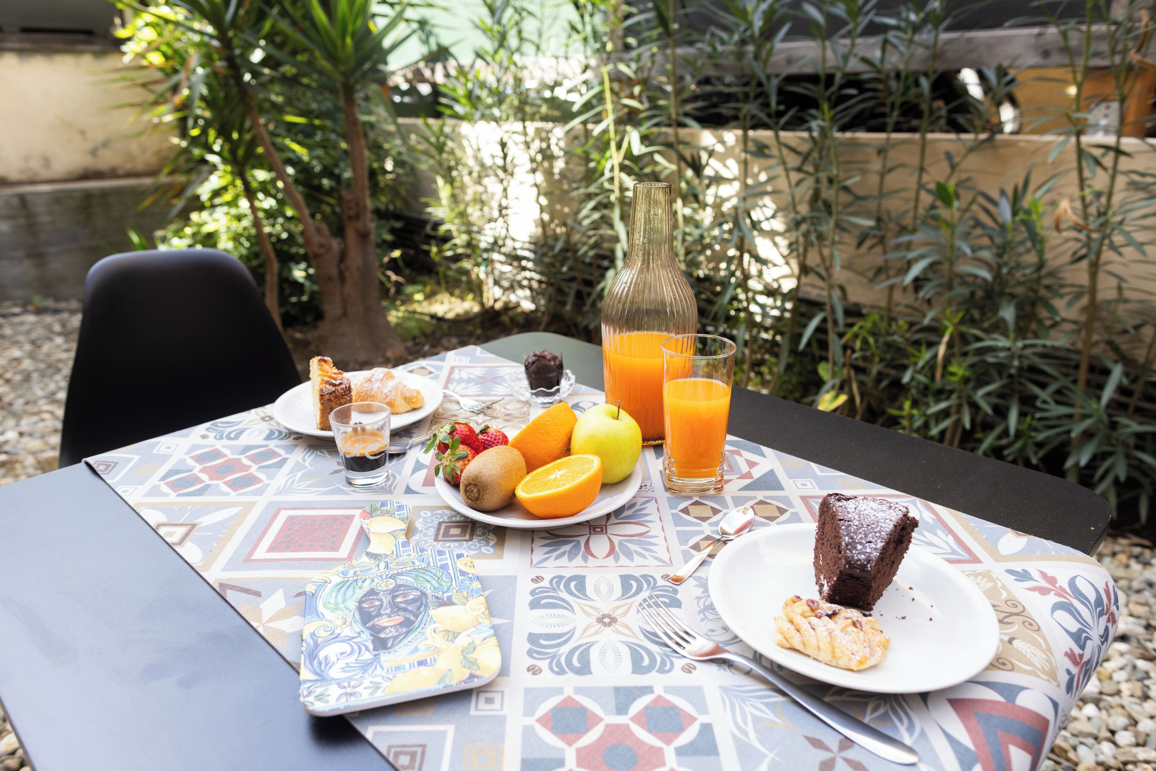 A table set with breakfast including fruits, juice, and a slice of cake, surrounded by greenery.