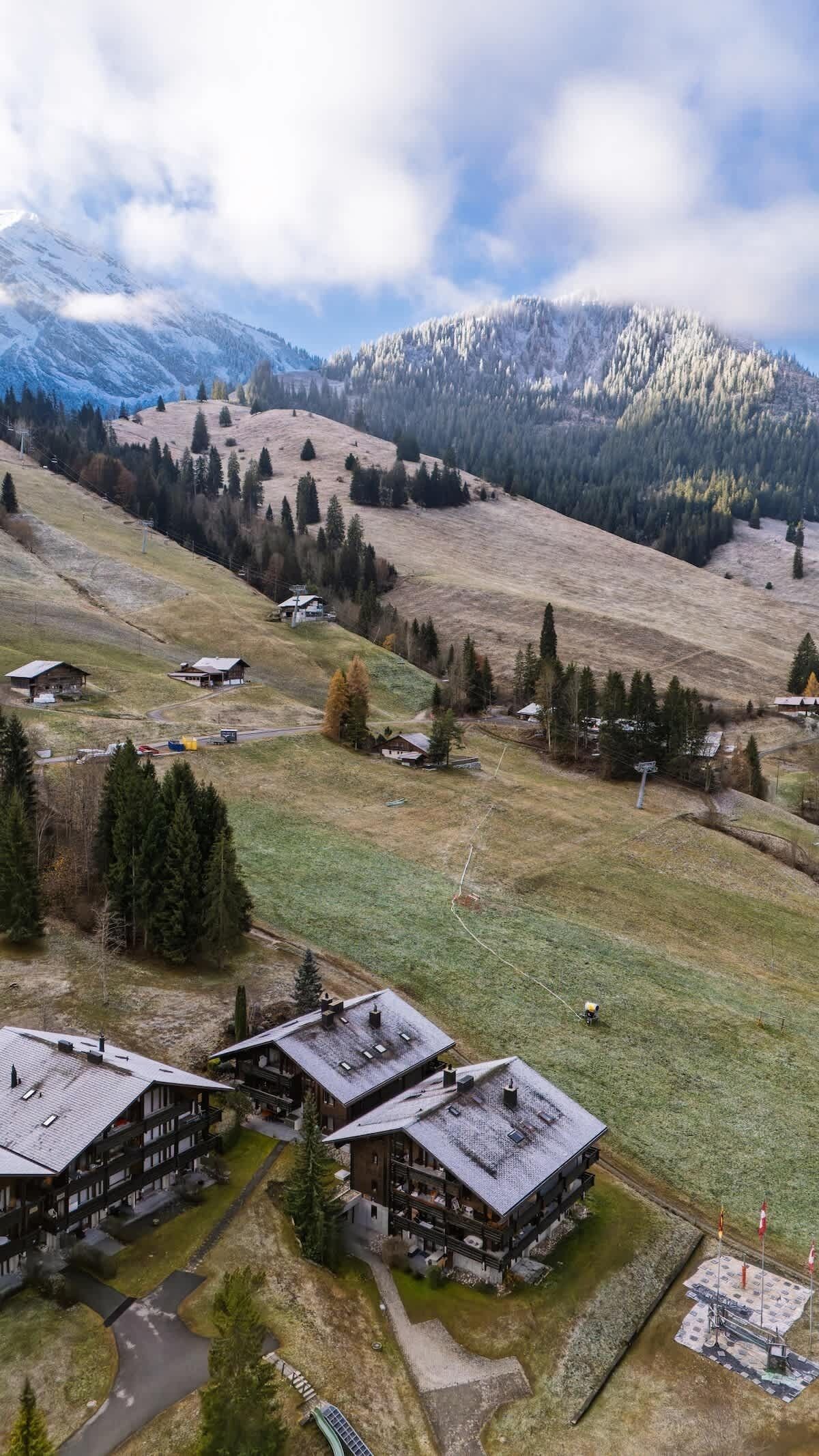 Un paisaje montañoso con un pequeño grupo de construcciones de madera, probablemente un albergue o complejo turístico, rodeado de árboles y un cielo despejado.