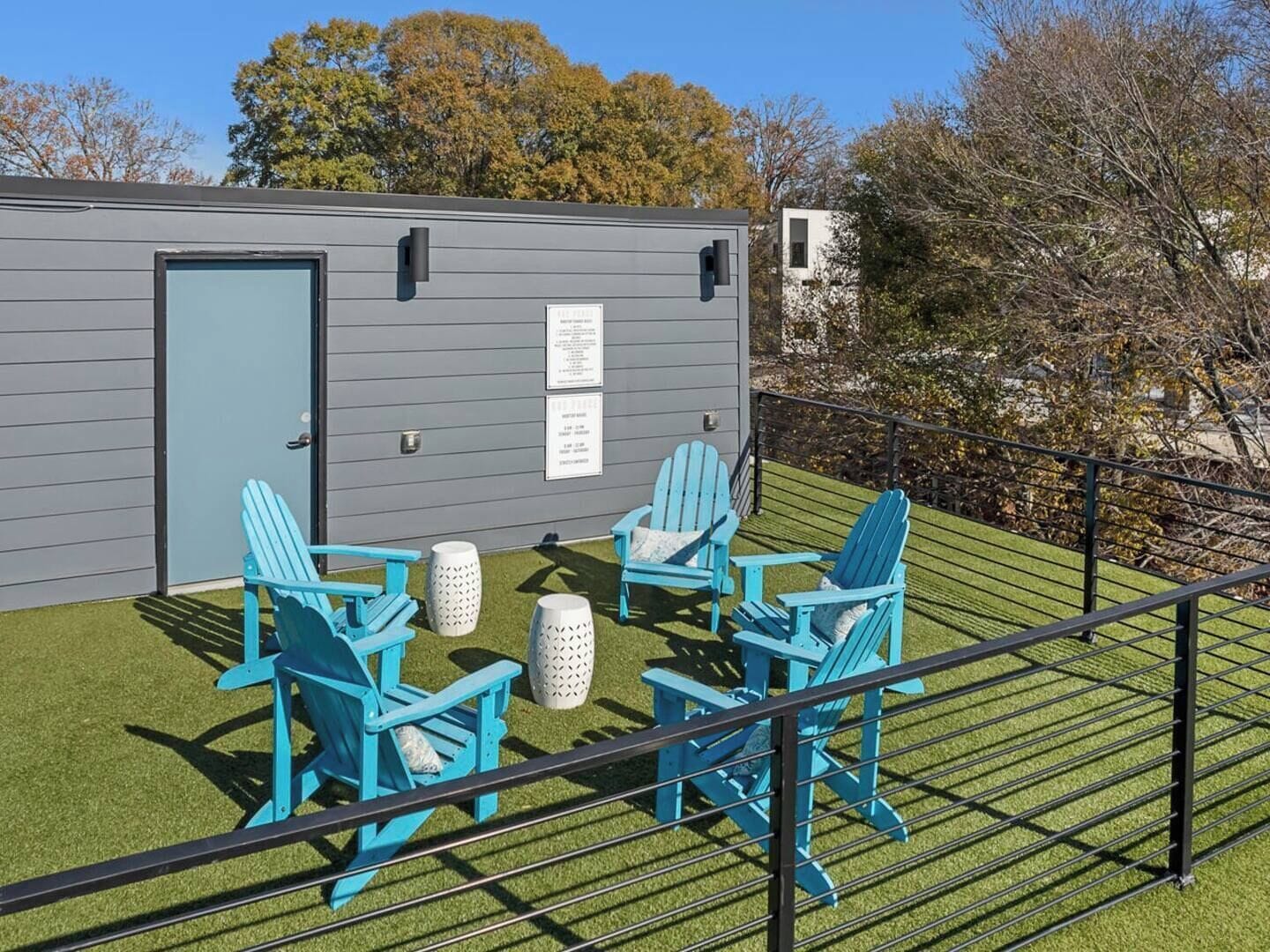 A small outdoor seating area with four blue Adirondack chairs and two white stools, a gray building with a door, and a black railing.
